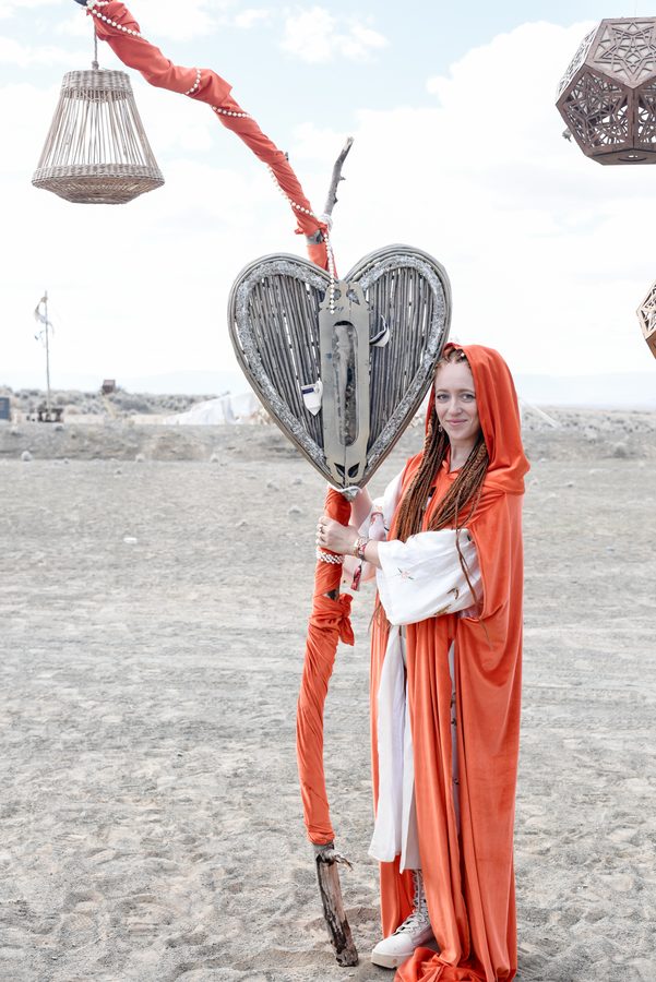 A figure in a red hooded robe holding a staff with a woven heart