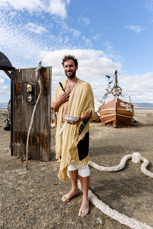 A camp mate in a yellow shawl holding a bowl at a wooden door